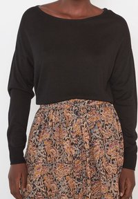 Pull en maille noire avec un col bateau et des manches longues et larges. Associé à une jupe à motifs multicolore ornée de motifs floraux.