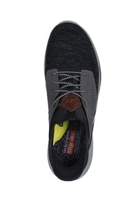 Zwarte en grijze Skechers instapschoen met een gestructureerde gebreide bovenkant, perforeerde grijze paneel en een bruin leren accent. Gele binnenzool zichtbaar.