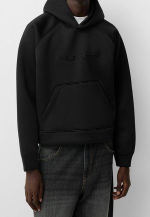 Schwarzer Hoodie mit Kängurutasche und dezent geprägtem "NUMÉ"-Logo, getragen mit dunkelgrauen Jeans an einer Person vor grauem Hintergrund.