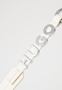 Cintura in pelle bianca con audace lettering metallico argento "HUGO". Texture liscia con finitura lucida e dettaglio con fibbia circolare.