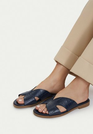 Füße tragen marineblaue offene Leder-Slide-Sandalen mit strukturiertem Muster und beige hochgekrempelte Hosen vor einfarbigem Hintergrund.