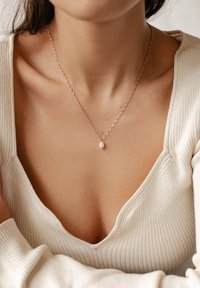 Roségoldkette mit einem einzelnen ovalen weißen Perlenanhänger, versehen mit einer zarten, gedrehten Kette, getragen über einem gerippten weißen Oberteil.