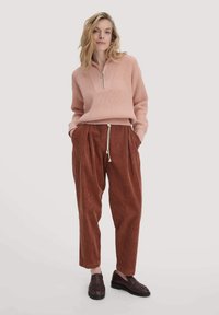 Helles rosa geripptes Sweatshirt mit Reißverschluss am Kragen, kombiniert mit lockeren, rostfarbigen Cordhose und dunkelbraunen Slip-On-Schuhen.