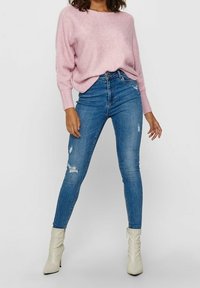 Pull en maille rose clair à coupe ample et texture côtelée, associé à un jean bleu ajusté avec des détails usés et des bottines blanches à la cheville.
