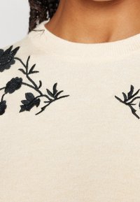 Pull beige avec broderie florale noire au niveau du col, présentant une texture côtelée et un design classique de col rond.