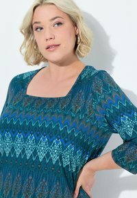 Tee in Türkis mit Muster, das über einen quadratischen Ausschnitt, Dreiviertelärmel und ein Zickzackdesign in Blau-, Grün- und Brauntönen verfügt.