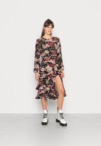 Vestido floral de mangas longas, base preta, padrões em rosa e bordô, bainha com babados, fenda lateral. Combinado com botas brancas com atacadores.