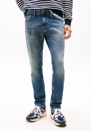 Mann trägt ausgewaschene blaue Slim-Fit-Jeans, marineblau-weiß gestreiften Pullover und marineblaue weiße Sneakers, steht vor weißem Hintergrund.