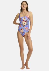 Une femme souriante se tient avec confiance dans un maillot de bain floral bleu, avec des bretelles et les pieds nus, sur un fond blanc simple.