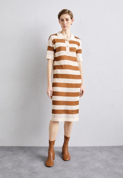 Robe en tricot avec des rayures horizontales marron et crème, manches courtes, col à revers et longueur mi-mollet. Portée avec des bottines marron.