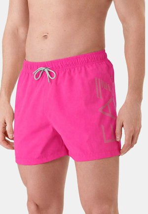 Uomo che indossa pantaloncini da bagno di un rosa acceso con coulisse grigia e una sottile scritta verticale sul lato destro, in piedi davanti a uno sfondo semplice.