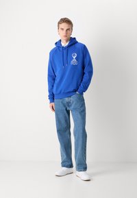 Sudadera azul con cordones y gráfico frontal, combinada con jeans de mezclilla azul claro y zapatillas blancas. Diseño simple con puños y borde de canalé.