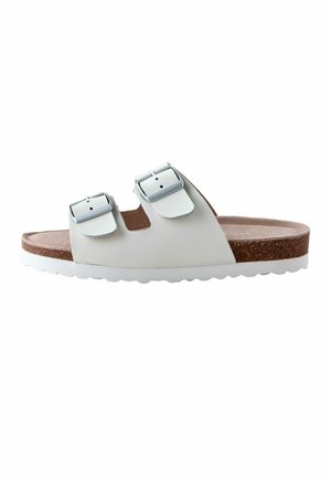 Witte slidesandalen met twee verstelbare zilveren gespen, een voetbed van kurk en een witte, gestructureerde zool, zijaanzicht op een witte achtergrond.