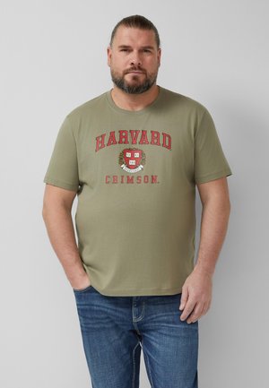 Man met baard die een olijfgroene Harvard Crimson T-shirt en een blauwe spijkerbroek draagt, staand tegen een effen lichtgrijze achtergrond.