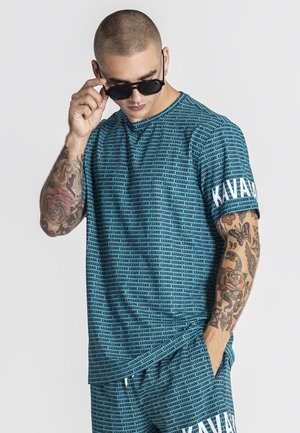 Uomo con tatuaggi che si sistema gli occhiali da sole neri, indossando una camicia e pantaloncini blu coordinati, entrambi coperti dal testo ripetuto "KAVANAGH GIANNI".