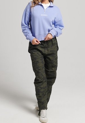 Kobieta idąca, ubrana w jasnofioletową bluzę z zamkiem do połowy długości i logo "Superdry", ciemnozielone moro spodnie typu cargo oraz białe trampki.
