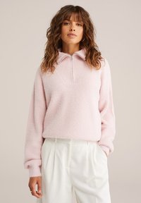 Gaiši rozā, teksturēts džemperis ar rāvējslēdzēju apkakli un garām piedurknēm, apvienots ar baltiem, krokotiem biksēm. Mīksta auduma ar ērtu dizainu.