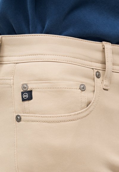 Κοντινό πλάνο τσέπης μπεζ παντελονιού με μεταλλικά τρουκς και μια μικρή μαύρη ετικέτα με το λογότυπο AG, φορεμένο με σκούρο μπλε πουκάμισο μέσα από το παντελόνι.
