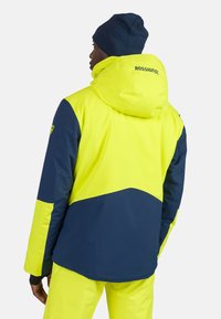 Felle gele en marineblauwe skijas met een capuchon, voorzien van een diagonale kleurblokontwerp en het logo op de achterkant. Glad, weersbestendig materiaal.