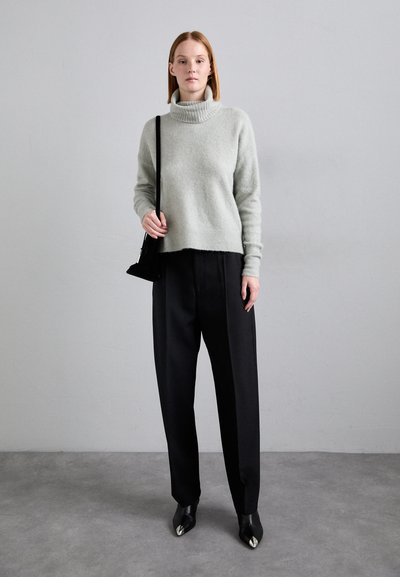 Maglione grigio chiaro a collo alto in materiale morbido, abbinato a pantaloni neri a vita alta. Il modello tiene una piccola borsa nera ed indossa scarpe con punta.