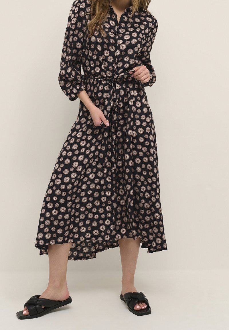 Femme portant une robe midi noire avec un motif floral circulaire léger, taille ceinturée et manches trois-quarts, associée à des sandales noires sans bride.