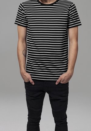 Man stands with hands in pockets wearing a black and white horizontal striped t-shirt and black pants against a gray background.