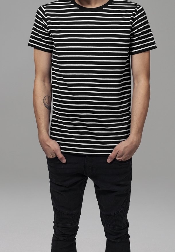 Un homme se tient les mains dans les poches, portant un t-shirt à rayures horizontales noires et blanches et un pantalon noir, sur un fond gris.
