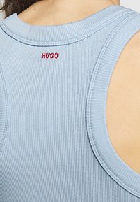 Top sin mangas azul con ribetes y la palabra HUGO bordada en rojo. Presenta amplios agujeros para los brazos y un escote redondo con detalles de costura visibles.