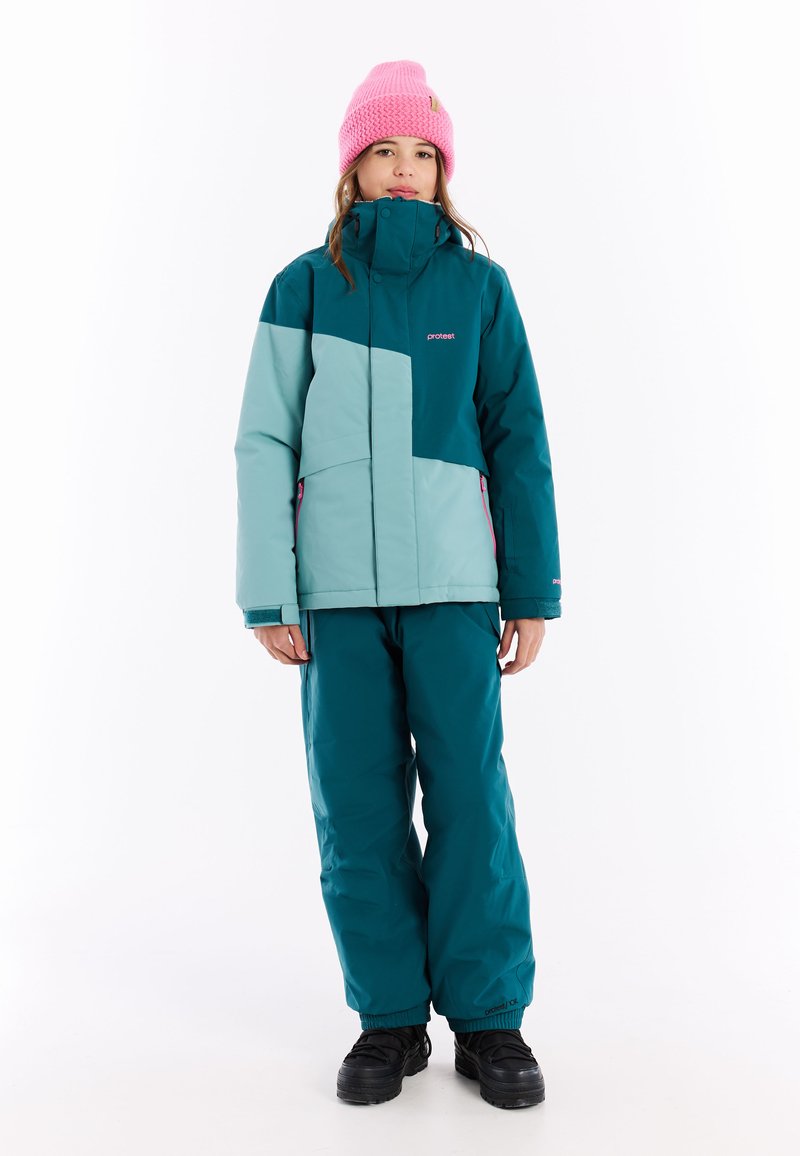 Tiefblau und hellblau gesteppte Schneejacke mit hohem Kragen, Reißverschlusstaschen und rosa Akzenten, kombiniert mit tiefblauen Schneehosen und schwarzen Stiefeln. Rosa Strickmütze.