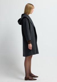 Grå uldfrakke med en stor hætte, oversized krave og sidelommer. Har lange ærmer og en skræddersyet silhuet. Brune fodtøj.