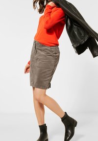 Jupe en velours côtelé kaki avec poches latérales, associée à un pull orange vif et une veste en cuir noir, complétée par des bottines noires.