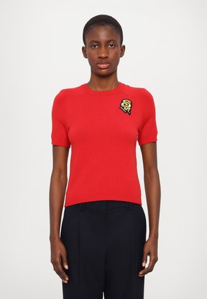 Pull en tricot rouge à manches courtes avec une silhouette ajustée. Présente un patch brodé noir et or sur la poitrine gauche. Texture lisse.