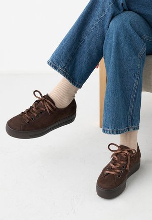 Person wearing beige socks, blue jeans, and dark brown suede sneakers sitting on a wooden chair with legs crossed.