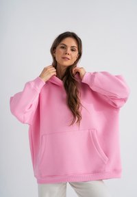 Felpa over-size rosa realizzata in materiale morbido, con tasca frontale e cappuccio con cordino; presenta polsini a coste e una vestibilità comoda.