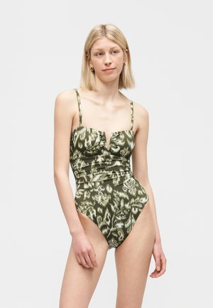 Ξανθιά γυναίκα που φοράει ένα πράσινο και μπεζ ολόσωμο μαγιό με λεπτούς ιμάντες στους ώμους, στέκεται μπροστά σε ένα απλό φωτεινό φόντο.