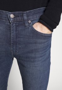 Jeans i denim i en mörkblå färg med en slät textur. Har en klassisk femfickdesign och en knappstängning fram.