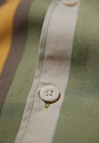 Detalle de camisa abotonada con rayas verticales en verde, beige y marrón. Presenta un botón redondo de color claro y costuras visibles.
