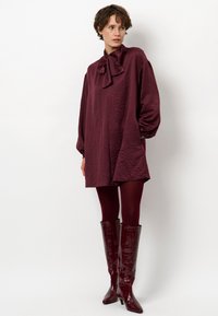 Bordeaux, gestructureerde jurk met een strikboordkraag, oversized mouwen en zijzakken, gecombineerd met glanzende kniehoge laarzen van krokodillenlederlook.