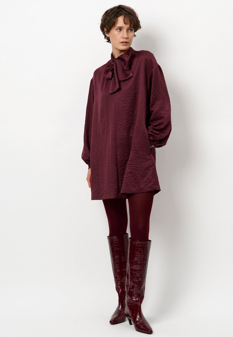 Bordeaux, gestructureerde jurk met een strikboordkraag, oversized mouwen en zijzakken, gecombineerd met glanzende kniehoge laarzen van krokodillenlederlook.