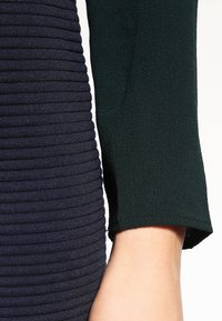 Haut côtelé bleu marine à manches longues avec une finition texturée, doté d'un revers de manche contrastant vert foncé. Tissu lisse, coupe ajustée.