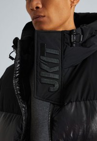 Giacca trapuntata nera con finitura lucida, collo grande con logo "JKIT" e dettagli con zip. Cappuccio attaccato per maggiore funzionalità.