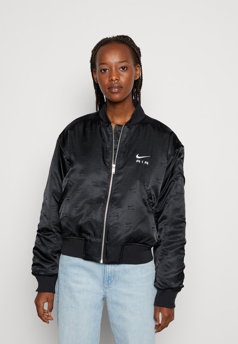 Giacca bomber nera in satin con zip, polsi e orlo a coste. Presenta loghi in rilievo e un design leggero. Indossata sopra jeans azzurri chiari.