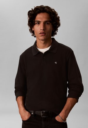Jeune homme aux cheveux bouclés porte un polo noir à manches longues, un tee-shirt blanc, une ceinture noire et un bracelet argenté, les mains dans les poches.