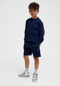 Marine sweatshirt en short set van zacht materiaal met logopatches. Model draagt grijs en witte sportschoenen met enkelkousen.
