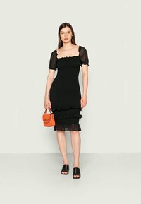 Vestido preto com mangas curtas bufantes, detalhes franzidos na bainha e na cintura, combinado com uma pequena mala laranja e sapatos slides pretos.