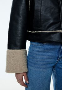 Schwarze Lederjacke mit beigen Lammfellmanschetten und -besatz, mit einem taillierten Design und glatter Textur. Kombiniert mit blauen Jeans.