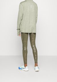 Jasnozielony sportowy polar z wysokim kołnierzem, w połączeniu z oliwkowymi legginsami z delikatnym wzorem oraz niebiesko-białymi butami do biegania.