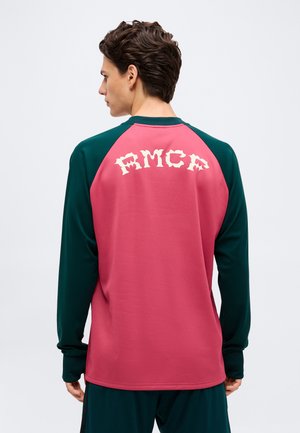 Mann mit dunklen Haaren, der ein langärmeliges pink-grünes Shirt mit „FMCP“ in weißer gotischer Schrift auf dem Rücken trägt, von der Kamera weg schauend.