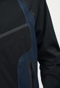 Μαύρο και ναυτικό μπουφάν με διαγώνια τσέπη με φερμουάρ και μικρό λογότυπο Nike στο στήθος, εμφανιζόμενο στον κορμό και το χέρι ενός ατόμου.