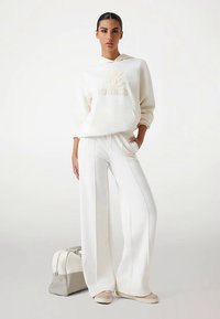 Weißes Sweatshirt mit einem geprägten "Guess"-Logo, kombiniert mit weißen Hosen mit weitem Bein. Eine hellfarbige Handtasche liegt neben dem Modell.